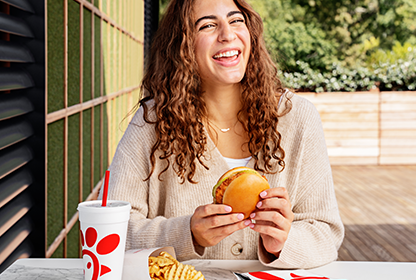A girl holding a Chick-fil-A Chicken Sandwich and smiling with Waffle Fries and a drink on the table