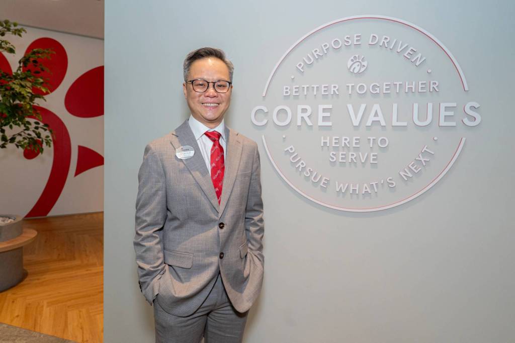 Chyn Koh, Singapore’s first local Owner-Operator, standing by a wall with the words "PURPOSE DRIVEN
BETTER TOGETHER
CORE VALUES
HERE TO SERVE
PURSUE WHAT'S NEXT"